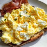 A slice of toast topped with creamy ricotta eggs and a side of bacon. This is Creamy Ricotta Egg Toast.