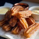 A pile of golden brown Easy Baked Churro Bites coated in cinnamon sugar, served with two dipping sauces.