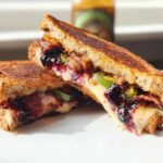 A gourmet blueberry grilled cheese sandwich, cut in half, showing melted cheese, blueberry jam, and jalapeno slices.