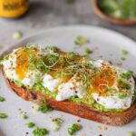 A slice of toast topped with mashed avocado, cottage cheese, poached eggs, sprouts, and green onions.
