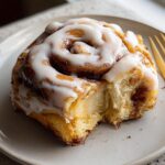 A bite taken out of a high-protein cinnamon roll bread, drizzled with icing, on a plate with a gold fork.
