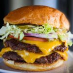 A tall, double-patty smash burger with melted cheddar cheese, fresh lettuce, tomato, onion, and pickles.