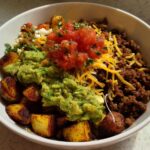 A close-up of a Loaded Potato Taco Bowl featuring roasted potatoes, seasoned ground beef, guacamole, salsa, and shredded cheese.