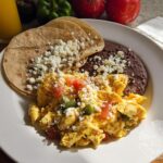 A plate of scrambled Mexican eggs with tomatoes and peppers, served with refried beans and crumbled cheese on tortillas.