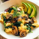 A plate of fluffy Mushroom Spinach Scrambled Eggs topped with fresh dill and sliced avocado.