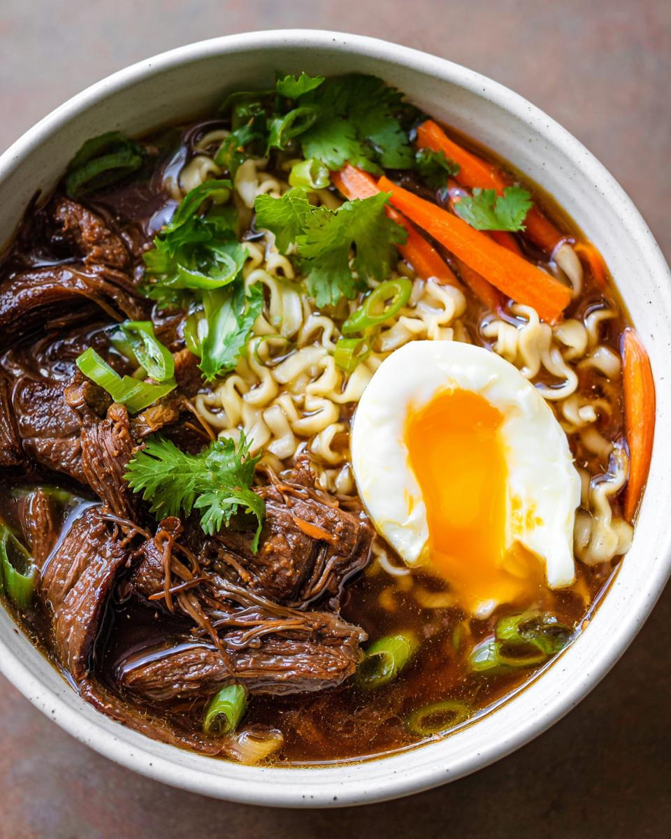 A close-up of a bowl of Slow Cooker Beef Ramen Noodles, featuring tender shredded beef, ramen noodles, sliced carrots, green onions, and a perfectly poached egg.