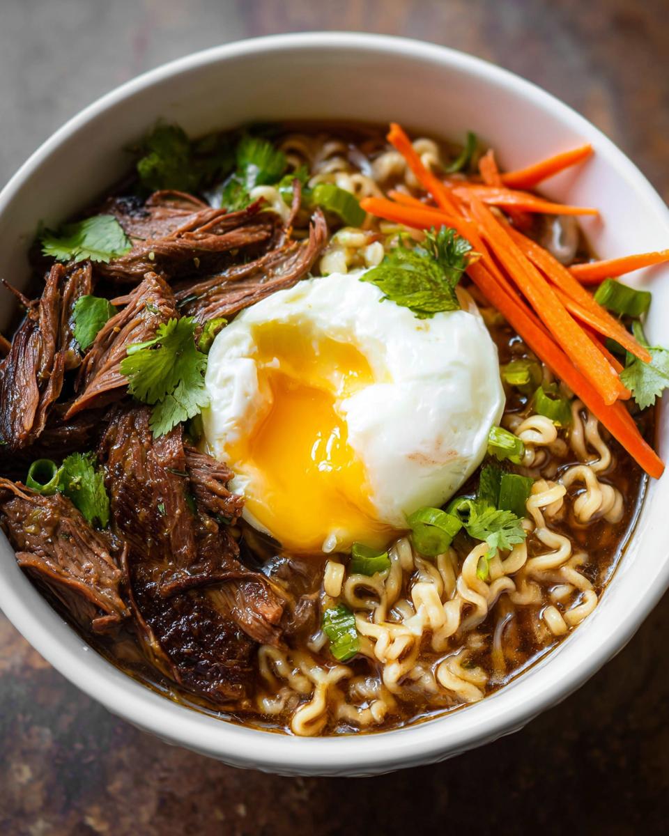 A close-up of a bowl of Slow Cooker Beef Ramen Noodles topped with shredded beef, a perfectly poached egg, shredded carrots, and green onions.