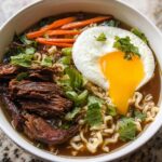 A steaming bowl of Slow Cooker Beef Ramen Noodles topped with a fried egg, shredded beef, carrots, and green onions.