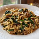 A close-up of a white bowl filled with creamy chicken sausage orzo pasta, featuring spinach and sun-dried tomatoes.