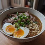 A bowl of Creamy Garlic Chicken Ramen topped with shredded chicken, green onions, sesame seeds, and a halved soft-boiled egg.