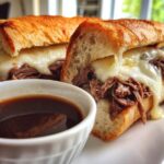 Close-up of a Crockpot French Dip Sandwich with melted cheese oozing over tender shredded beef on a toasted roll, served with au jus.