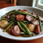 A close-up of a white bowl filled with Crockpot Kielbasa and Green Beans, featuring sliced kielbasa, whole baby potatoes, and green beans in a savory sauce.