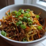 A close-up of a white bowl filled with Egg Roll in a Bowl, featuring ground meat, shredded cabbage, carrots, and topped with fresh green onions.