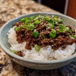 A close-up of a Korean beef rice bowl, featuring seasoned ground beef over white rice, topped with green onions and sesame seeds.