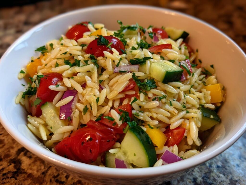 A close-up of a bowl filled with lemony orzo salad with fresh vegetables like tomatoes, cucumber, red onion, and yellow bell pepper.