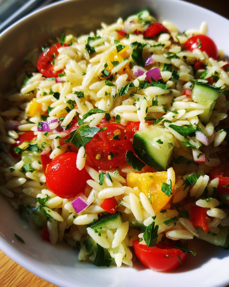 Close-up of a vibrant lemony orzo salad with fresh veggies like cherry tomatoes, cucumber, red onion, and parsley.