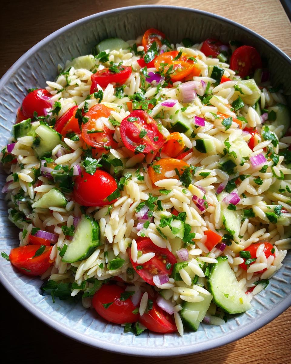 A close-up of a bowl filled with lemony orzo salad, featuring cherry tomatoes, cucumber, red onion, and parsley.