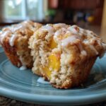 Close-up of a moist peach muffin, split open to reveal juicy peach chunks and drizzled with vanilla glaze.