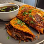 Close-up of My Fave Birria Tacos on a gray plate, served with a side of consommé.