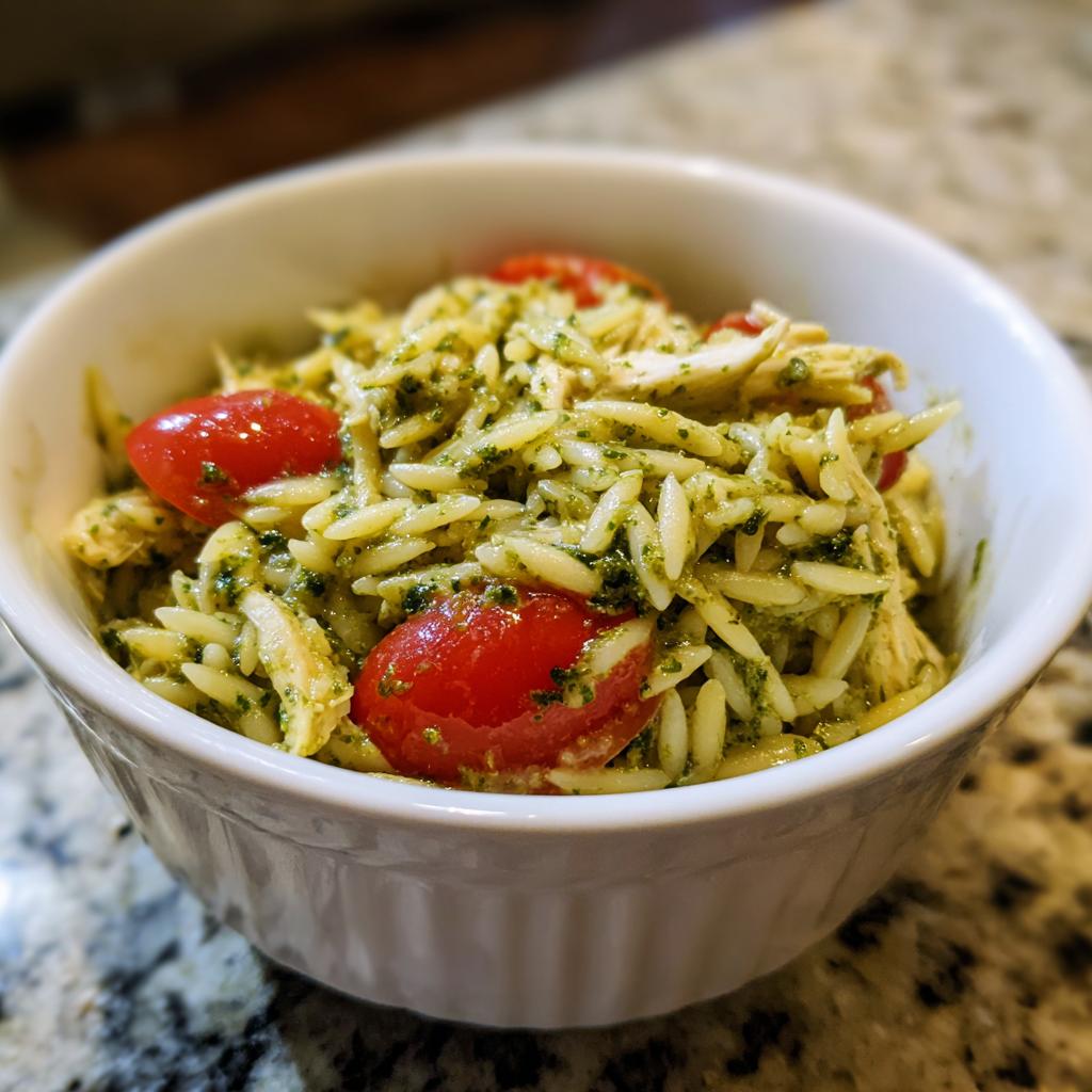A bowl of pesto orzo pasta with shredded chicken and halved cherry tomatoes, a vibrant and flavorful dish.