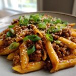 A close-up of a bowl of Sticky Honey Garlic Sausage Pasta Skillet, garnished with green onions and sesame seeds.